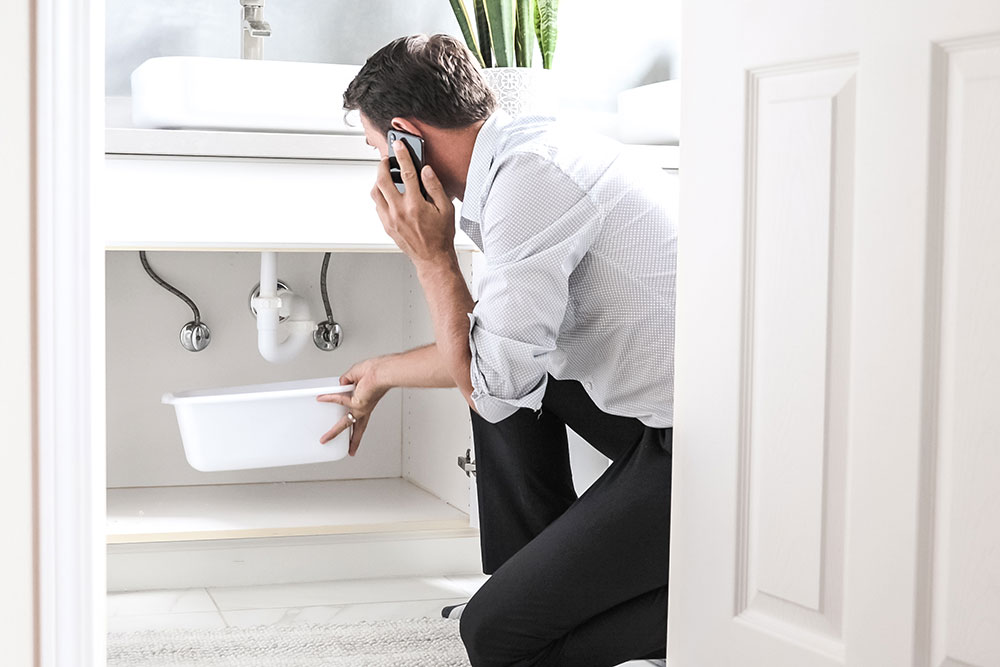 Man on phone fixing sink leak with bucket under pipes.