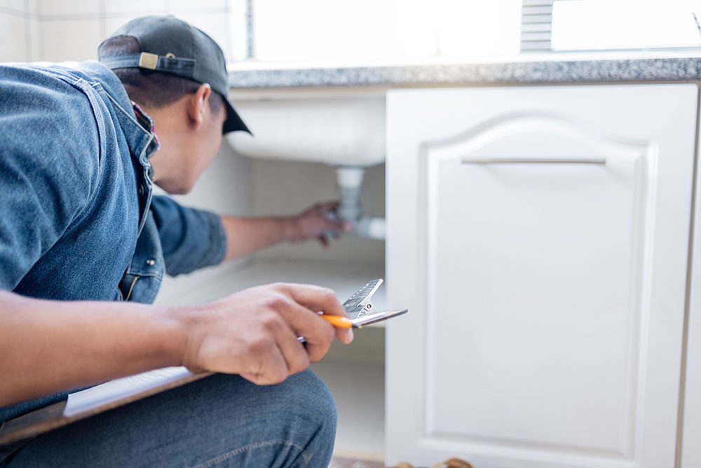 Plumber inspecting sink pipes with tool in hand, focusing on kitchen plumbing repair.