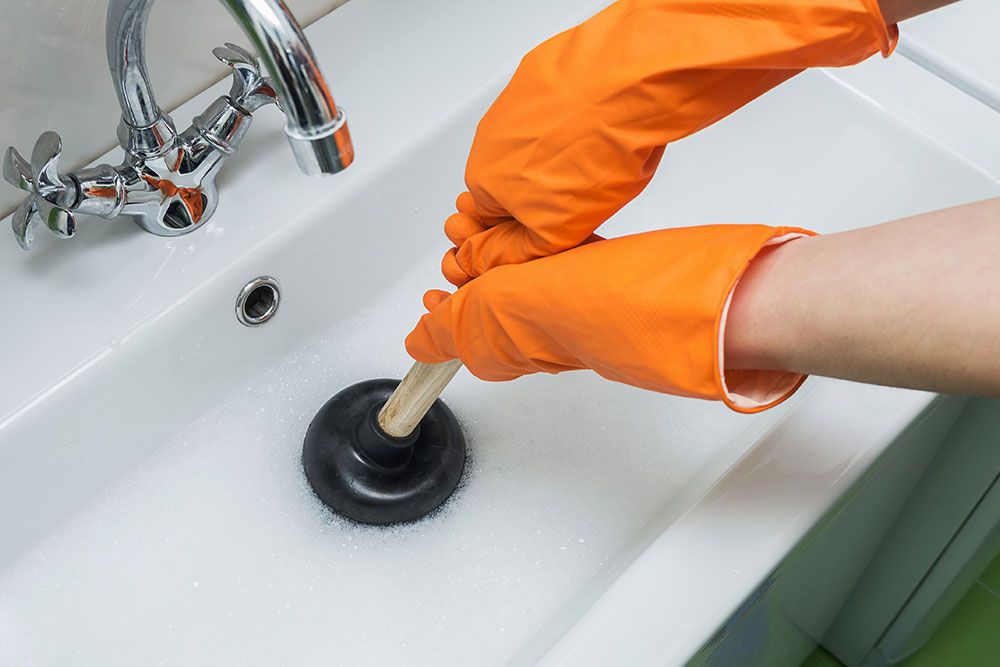Person unclogging bathroom sink with plunger and wearing orange gloves for effective home plumbing maintenance.